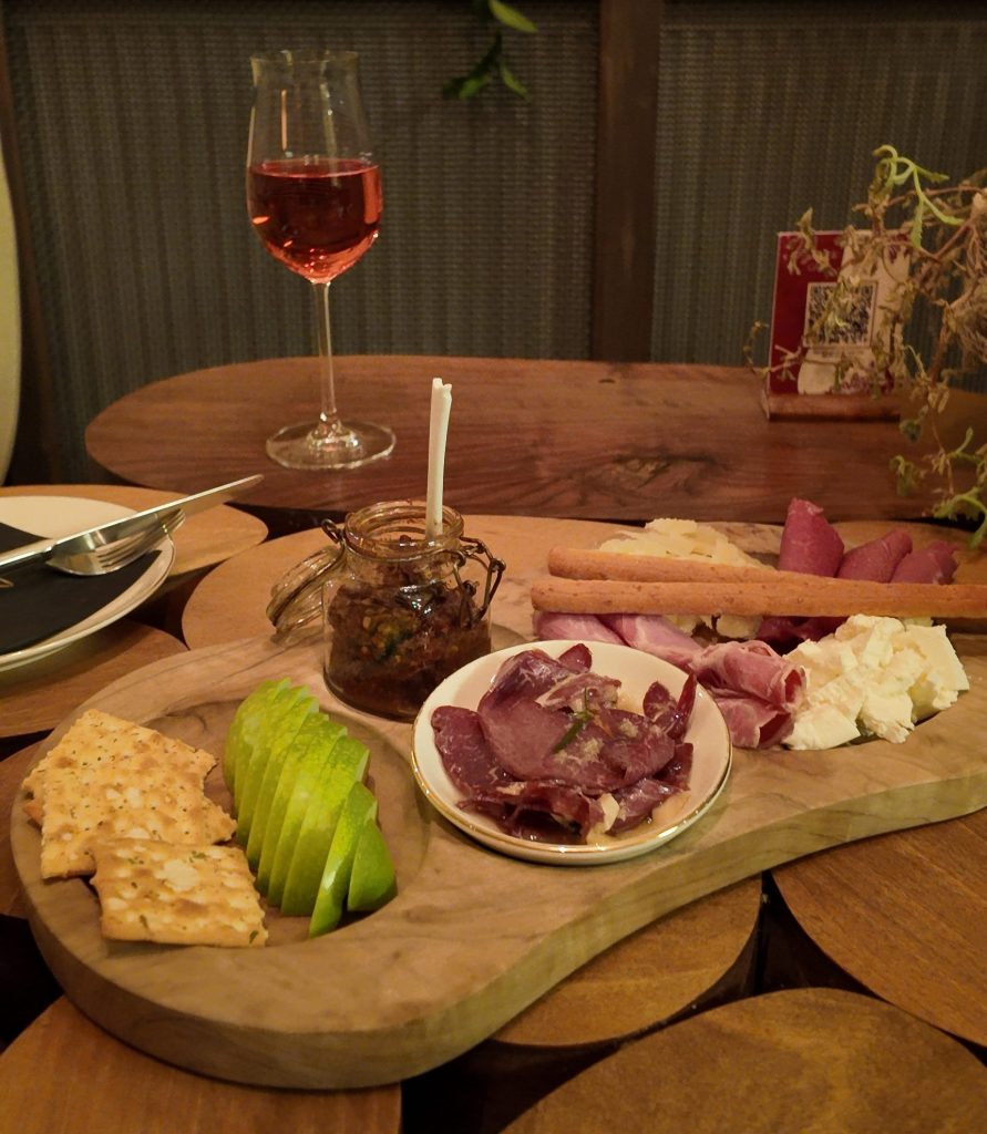 A wooden serving board featuring slices of green apple, assorted cheeses, cured meats, and crackers, accompanied by a small jar of spread and a glass of rosé wine.